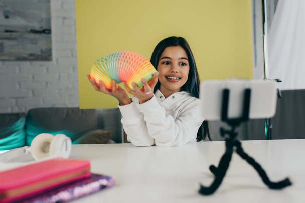 vrolijk tiener meisje tonen spiraal speelgoed in de buurt wazig smartphone en gele achtergrond thuis - Foto, afbeelding