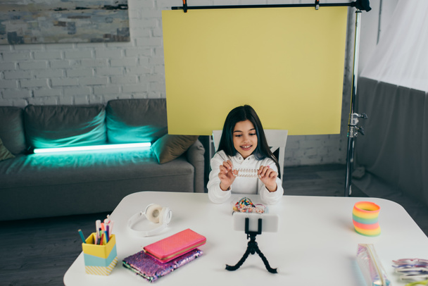 χαμογελαστό κορίτσι κρατώντας βραχιόλι χάντρες, ενώ καταγραφή βίντεο blog στο smartphone στο σπίτι - Φωτογραφία, εικόνα