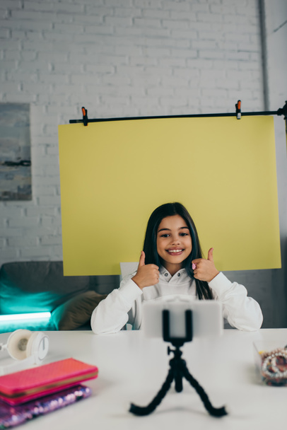 счастливый видео блоггер показывает большие пальцы вверх на желтом фоне рядом с мобильным телефоном дома - Фото, изображение