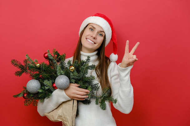 Portret van gelukkige mooie jonge vrouw in Santa Claus hoed met boeket van sparren takken tonen overwinning teken, poseren geïsoleerd over rode kleur achtergrond muur in de studio. Nieuwjaar 2023 vakantie concept - Foto, afbeelding