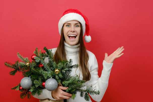 Shocked young Santa woman 20s wears Christmas hat holding bouquet of spruce branches raised hand, posing isolated on red color background wall in studio. Happy New Year 2023 celebration concept - Foto, Bild