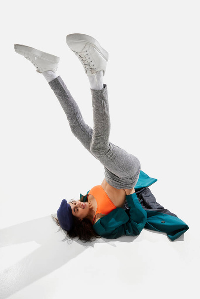 Portrait of young woman in green trench coat lying on floor with legs up, posing over white background. Concept of beauty, modern fashion, extraordinary style, self-love, self-acceptance, uniqueness - Foto, afbeelding