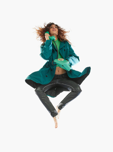 Portrait of young woman in green trench coat jumping, talking on vintage phone over white background. Concept of beauty, modern fashion, extraordinary style, self-love, self-acceptance, uniqueness - Φωτογραφία, εικόνα