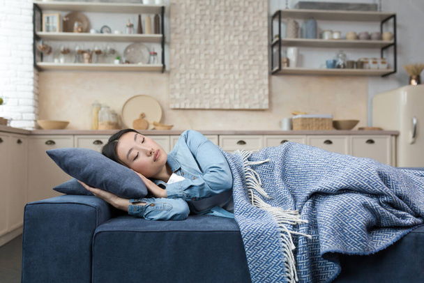 Close-up photo of a young beautiful Asian woman sleeping at home lying on the sofa covered with a blanket, resting after work for in the living room woman with closed eyes. - Foto, imagen