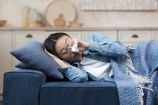 Close-up photo of a sick woman lying on the sofa at home, having a fever and flu, an Asian woman lying under a blanket on the sofa. - Fotoğraf, Görsel