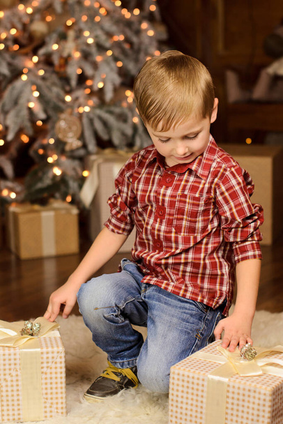 portrait of a blond boy in a red plaid shirt, sitting near the Christmas tree and boxes with gifts, considering them - Foto, afbeelding