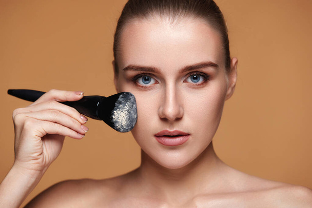 beautiful young woman applying powder with professional make-up brush on beige background - Photo, image