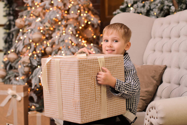 pieni vaalea poika hymyilee, pitää suurta laatikkoa, jolla on lahja uudelle vuodelle käsissään, istuu kevyessä tuolissa - Valokuva, kuva