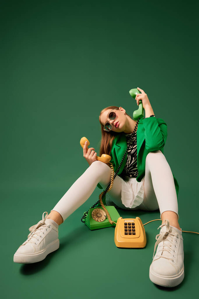 Creative portrait of young without emotions girl in green jacket sitting on chair with retro phone isolated over green background. Vivid style, beauty, queer, freak, fashion concept. Copy space for ad - Фото, зображення