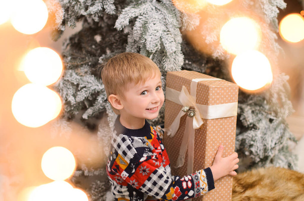 Pieni poika villapaidassa lumen peitossa pitää lahjapakkausta kädessään ja hymyilee. Ammuttu seppeleen valoissa, bokeh - Valokuva, kuva
