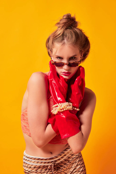 Look. Half-length portrait of young fashionable girl in red huge latex glove and sunglasses isolated over bright yellow background. Vivid style, queer, art, fashion. Weird people concept. - Φωτογραφία, εικόνα