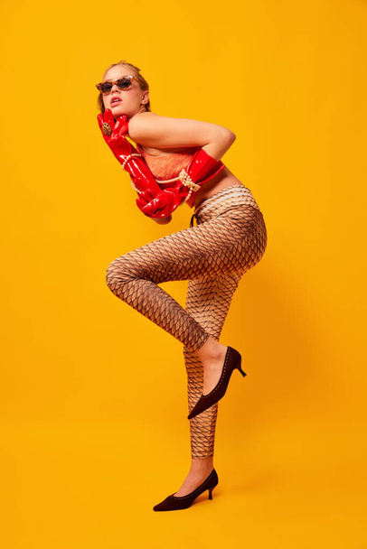 Aspiration. Weird people concept. Creative portrait of young girl in avant-garde style image isolated on bright yellow background. Vivid style, queer, art, fashion Stylish model in bright red gloves. - Photo, Image