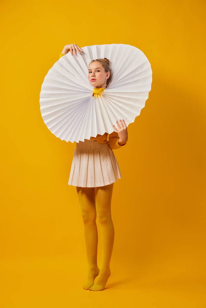 Sunny. Young indifferent girl in giant jabot collar or neckwear and yellow tights isolated over yellow background. Contemporary art, weird beauty, avant-garde fashion. - Foto, Imagen
