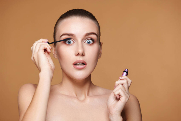 surprised woman applying black mascara on long thick eyelashes with brush and making makeup on beige background - Photo, image