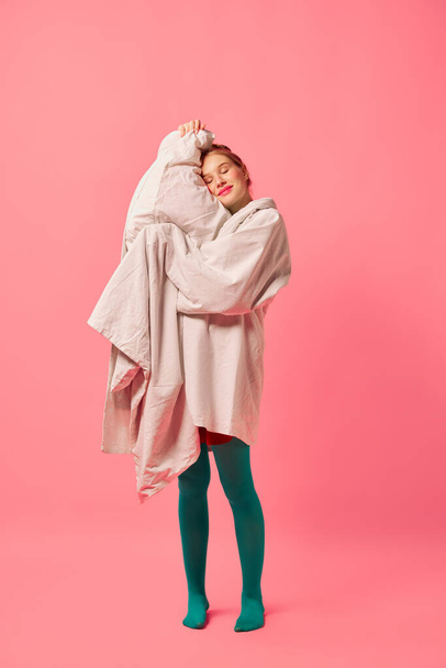 Beautiful young girl wrapped in soft blanket standing isolated over pink background. Comfrort, fashion, art, creativity. Weird people concept. Copy space for ad - Photo, image