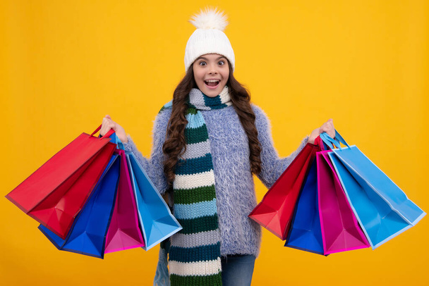 Teenager child girl in winter hat with shopping bags on yellow background. Kid with shopping winter sale bags. Excited teenager girl - Valokuva, kuva