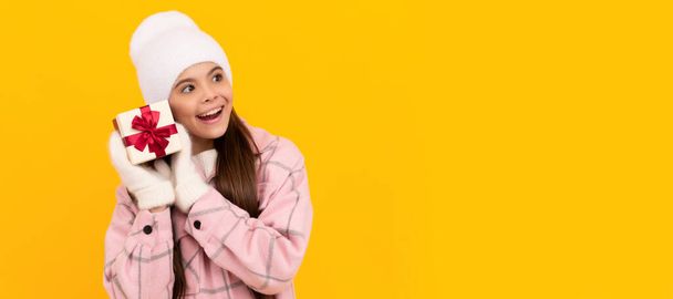 curious kid in hat and mittens hold gift box on yellow background, new year. Banner of christmas child girl, studio kid winter portrait with copy space - Photo, Image