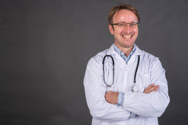 Médico varón confiado sonriendo con los brazos cruzados sobre fondo gris plano horizontal con espacio de copia - Foto, Imagen
