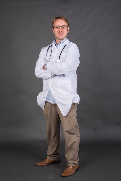 Tiro de cuerpo entero de médico masculino con estetoscopio sobre el cuello con abrigo médico contra fondo gris tiro vertical - Foto, imagen