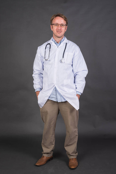 Tiro de cuerpo entero de médico masculino con estetoscopio sobre el cuello con abrigo médico contra fondo gris tiro vertical - Foto, imagen