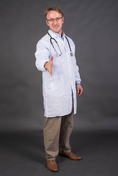 Médico masculino sonriente con estetoscopio sobre el cuello en abrigo médico que ofrece apretón de manos contra fondo gris tiro vertical - Foto, imagen