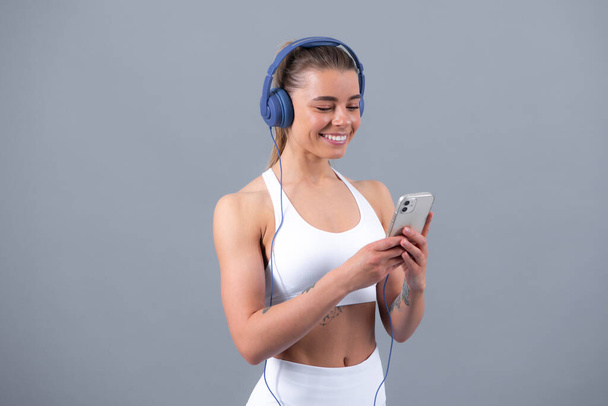 Sporty girl with headphone choose music for sport. Full body of young cheerful smiling woman in sports wear, isolated over gray background. Healthy beautiful sportswoman - Photo, image