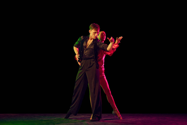 Elegance. Full-length portrait of young beautiful man and woman dancing ballroom dance isolated over dark background in neon light. Concept of art, beauty, grace, action, emotions. - Foto, afbeelding