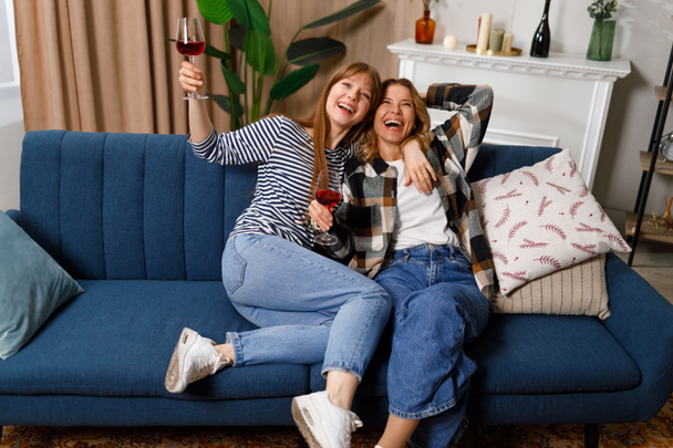 Cheerful mature mother and adult daughter talking, laughing and hugging while drinking wine on sofa at home - Foto, Bild
