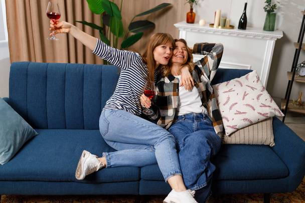 Allegro madre matura e figlia adulta parlando, ridendo e abbracciandosi mentre beve vino sul divano di casa - Foto, immagini