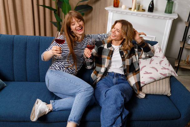 Dwie kobiety w różnym wieku piją wino i dobrze się bawią siedząc na kanapie - Zdjęcie, obraz