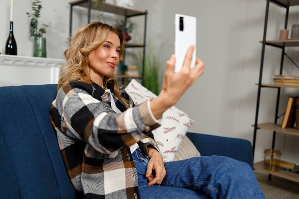 Donna attraente in posa sulla fotocamera dello smartphone mentre seduto sul divano in appartamento - Foto, immagini