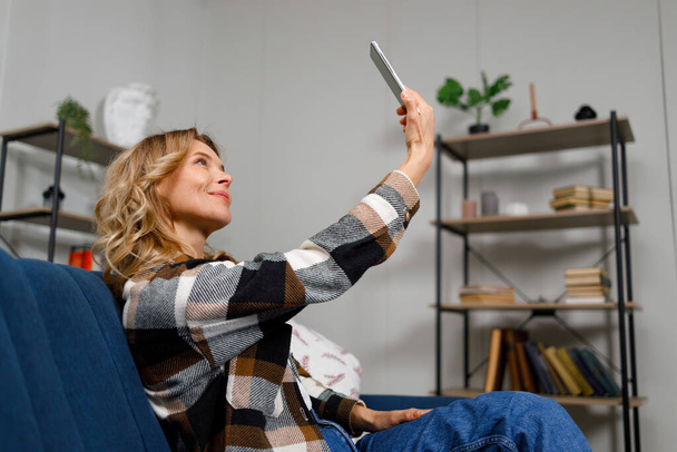 Mulher loira tomando selfie no telefone celular enquanto sentada no sofá na sala de estar - Foto, Imagem