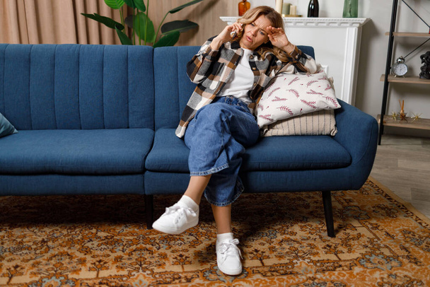 Järkyttynyt nainen puhuu puhelimessa istuessaan sohvalla - Valokuva, kuva