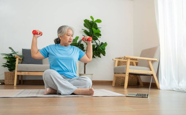 Азиатская старшая женщина поднимает гантель для упражнений и тренировок дома. Активная зрелая женщина делает упражнения на растяжку в гостиной. Активные и здоровые упражнения для пожилых, пожилых и пожилых людей. - Фото, изображение