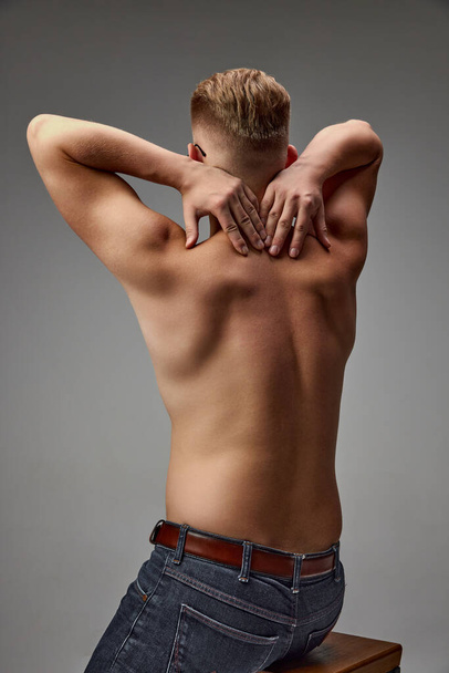 Portret van jonge gespierde man die shirtloos geïsoleerd poseert over een grijze studio achtergrond. Terugmasseren om pijn te verminderen. Concept van de gezondheid van de mens, natuurlijke schoonheid, lichaams- en huidverzorging, geneeskunde - Foto, afbeelding