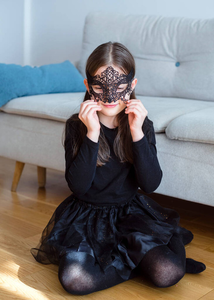 カーニバルの仮面と黒の衣装を着た7歳の少女の肖像画。接近中だ。ハロウィン・コンプ - 写真・画像
