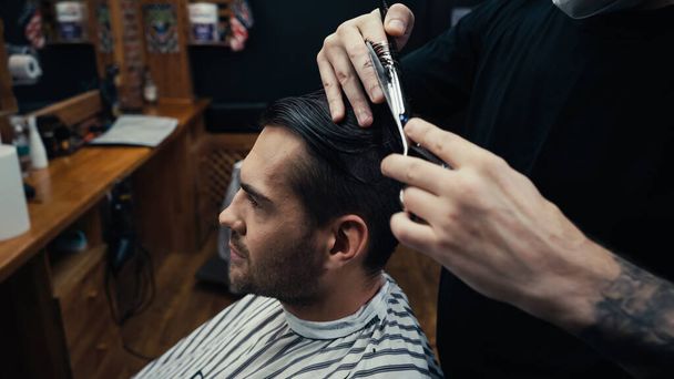 Barber cutting hair of customer in hairdressing cape in barbershop  - Photo, Image
