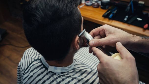 Barber trimming hair of man in hairdressing cape in blurred barbershop  - Photo, Image