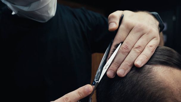 Обрезанный вид парикмахера в медицинской маске, стригущего волосы человека в парикмахерской  - Фото, изображение