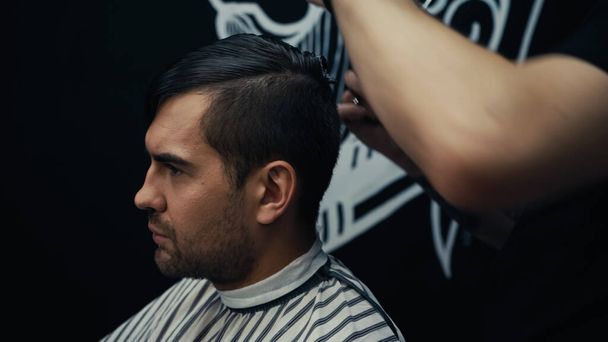 Hombre en capa de peluquería sentado cerca de estilista en la barbería  - Foto, Imagen