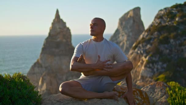 Handsome man performing yoga exercise in morning sunlight beautiful ocean rock. Focused strong athlete relax after workout at stunning nature. Serene guy rest at seashore. Zen like lifestyle concept - Footage, Video