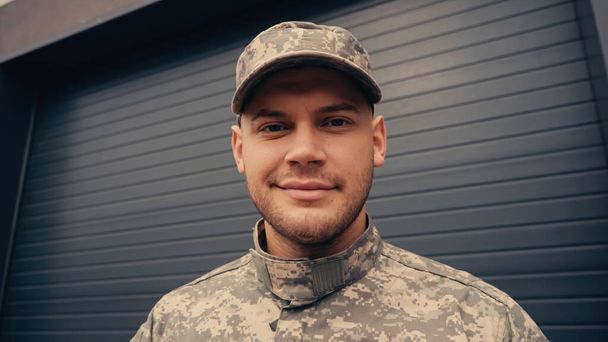 soldado en uniforme y gorra sonriendo mientras mira la cámara cerca del edificio  - Foto, Imagen