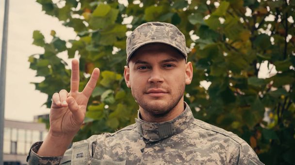 Üniformalı ve şapkalı genç asker ağaçların yanında barış işareti gösteriyor. - Fotoğraf, Görsel