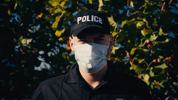 πορτρέτο του νεαρού αστυνομικού με στολή και ιατρική μάσκα που στέκεται κοντά στο δέντρο  - Φωτογραφία, εικόνα