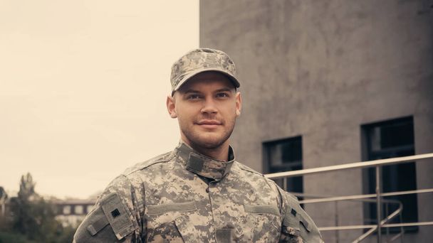 Soldado sonriente en uniforme y gorra mirando hacia otro lado mientras camina al aire libre  - Foto, Imagen