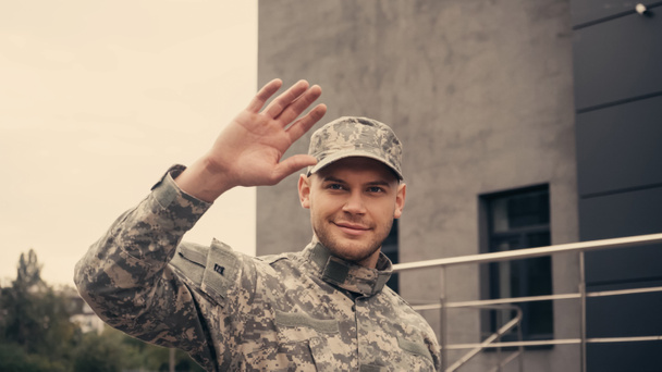 Soldado sorridente em uniforme e boné cumprimentando enquanto caminhava ao ar livre  - Foto, Imagem