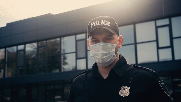 oficial de policía en máscara médica y gorra con letras mirando a la cámara cerca del edificio  - Foto, Imagen