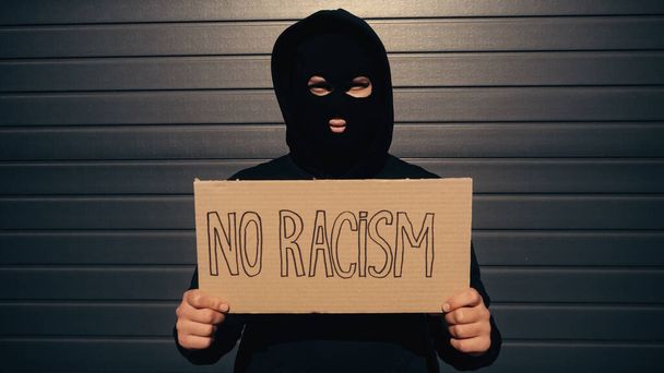 Mann mit Sturmhaube hält Plakat mit rassistischem Schriftzug in Hausnähe  - Foto, Bild