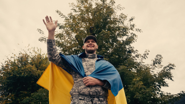vista de ángulo bajo de soldado sonriente en uniforme militar agitando la mano mientras sostiene la bandera ucraniana al aire libre  - Foto, Imagen