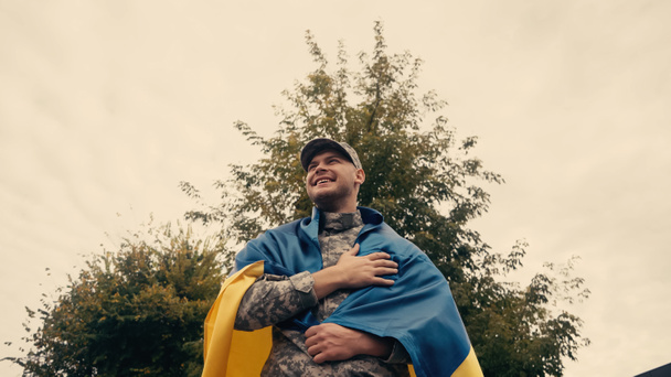χαμηλή γωνία άποψη του χαρούμενος και περήφανος στρατιώτης με στρατιωτική στολή κρατώντας ουκρανική σημαία σε εξωτερικούς χώρους  - Φωτογραφία, εικόνα